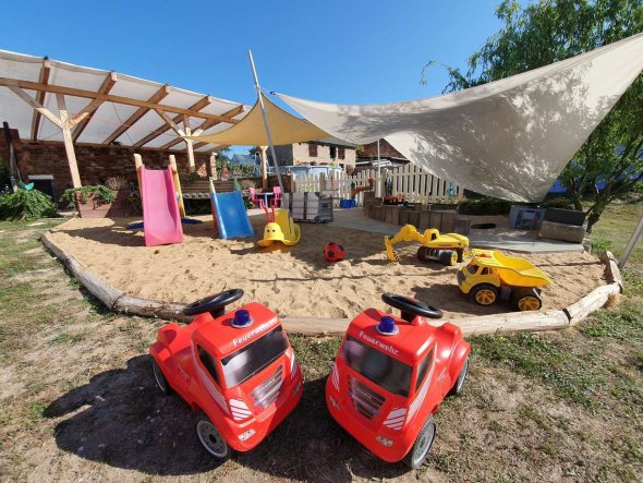 Ein großer Sandkasten über dem Sonnensegel gespannt sind. Im Sandkasten sind verschiedene Spielzeuge wie z.B. Autos, Bälle oder Rutschen zu sehen.